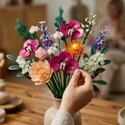 A close-up of a Mavirea building block floral arrangement resting on a rustic wooden table in a warm, sunlit room. The detailed textures of the orange gerberas and lush green stems emphasize its high-quality design as a maintenance-free interior accent.