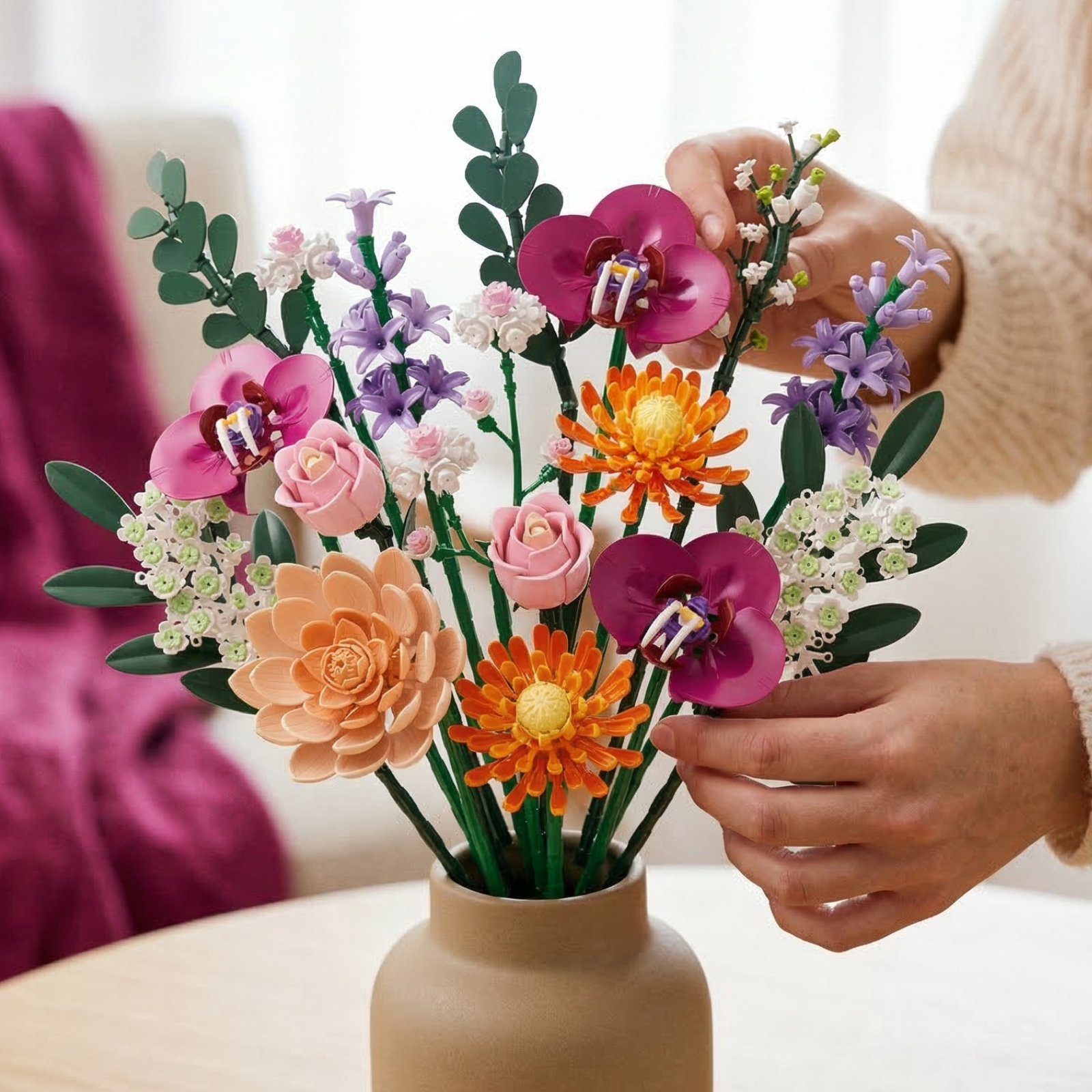 A hand carefully arranging a full Mavirea building block flower bouquet in a chic ceramic vase. The arrangement features vibrant pink roses, purple hyacinths, and deep magenta orchids, showcasing its use as a sophisticated, everlasting home decor centerpiece.