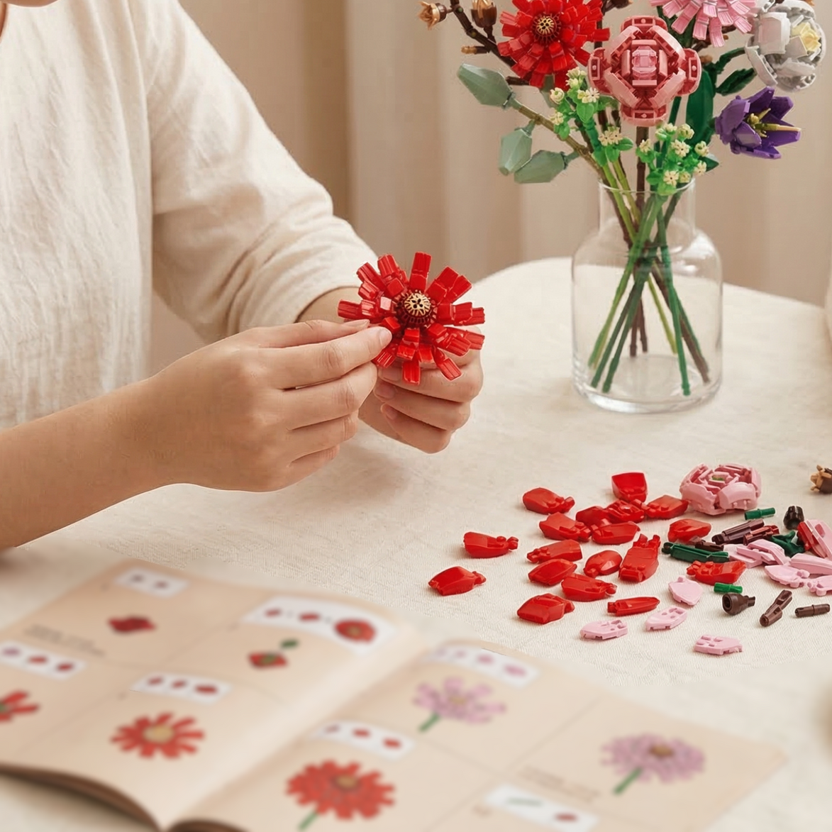 A person assembling a vibrant red Mavirea building block flower following an instruction manual, with a completed colorful bouquet in a glass vase in the background.
