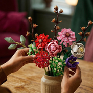 Close-up of hands arranging a diverse Mavirea floral bouquet—featuring red, pink, and purple building block blooms—into a ceramic vase for an elegant, eternal home display.