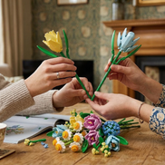 Two people holding up completed yellow and blue Mavirea flower stems over a vibrant pile of building block blooms, showcasing a shared moment of creating an eternal gift.