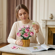 A woman thoughtfully assembles the Mavirea Pink Rose Potted Plant building kit at a marble table with the instruction manual open, highlighting it as an engaging and creative DIY gift.